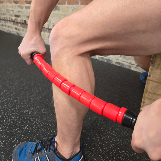 A person using the White Lion Athletics 23.5" 'Reflex' Flexible Massage Stick on their lower leg, focusing on relief for the calf muscle in a gym setting.