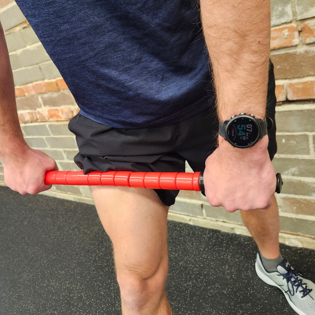 Male athlete using White Lion Athletics Big Red Massage Stick on their left quad while standing in a gym.