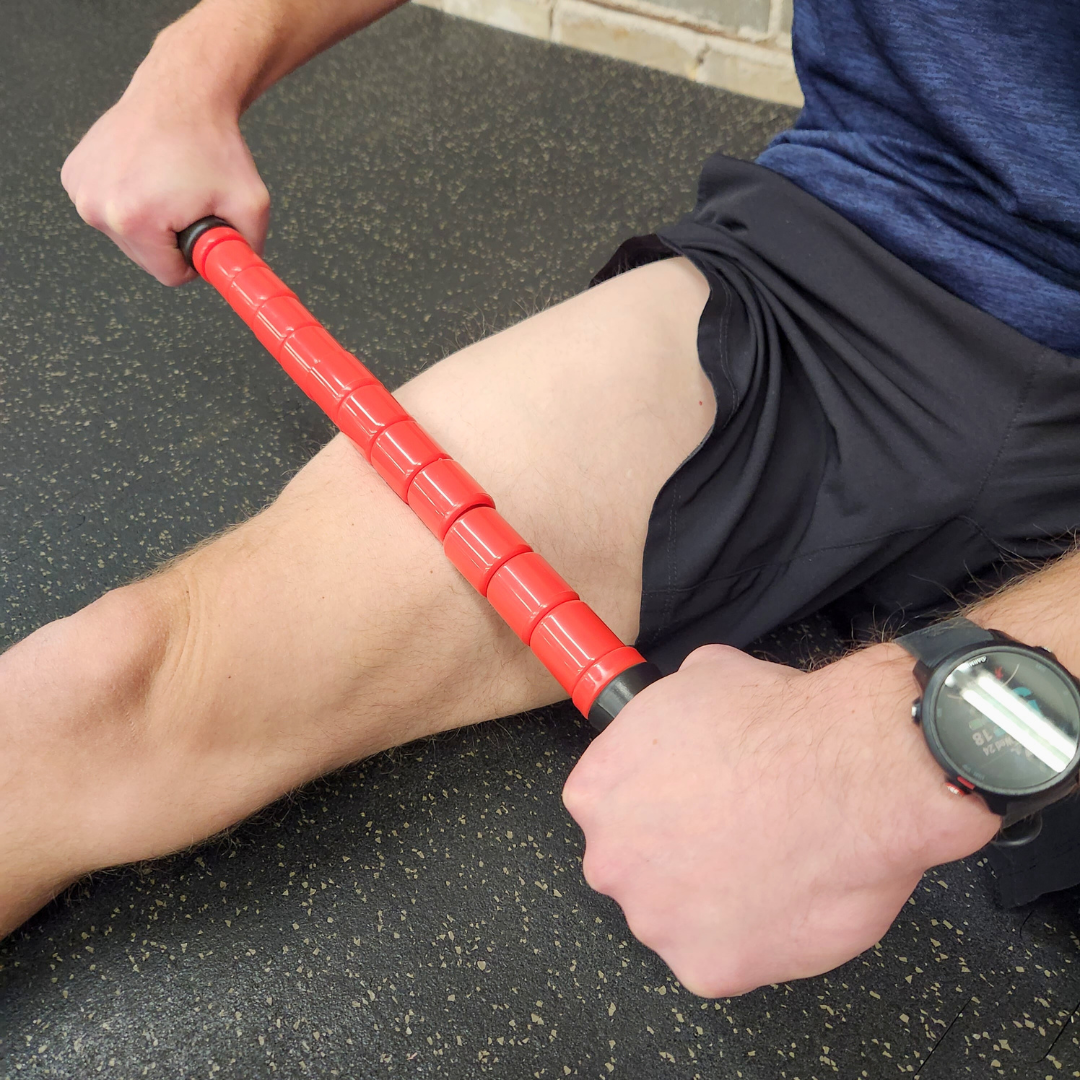 Male athlete using White Lion Athletics Big Red Massage Stick on their right quadricep while sitting on a gym floor.