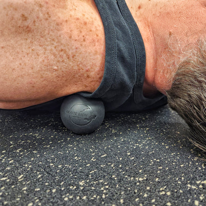 Man laying on his back with a black, White Lion Athletics lacrosse ball with a laser engraved logo on his left trapezius muscle.