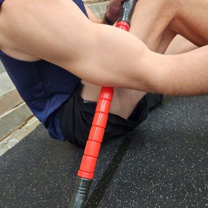 Male athlete using White Lion Athletics Big Red Massage Stick on their biceps while sitting on the gym floor.