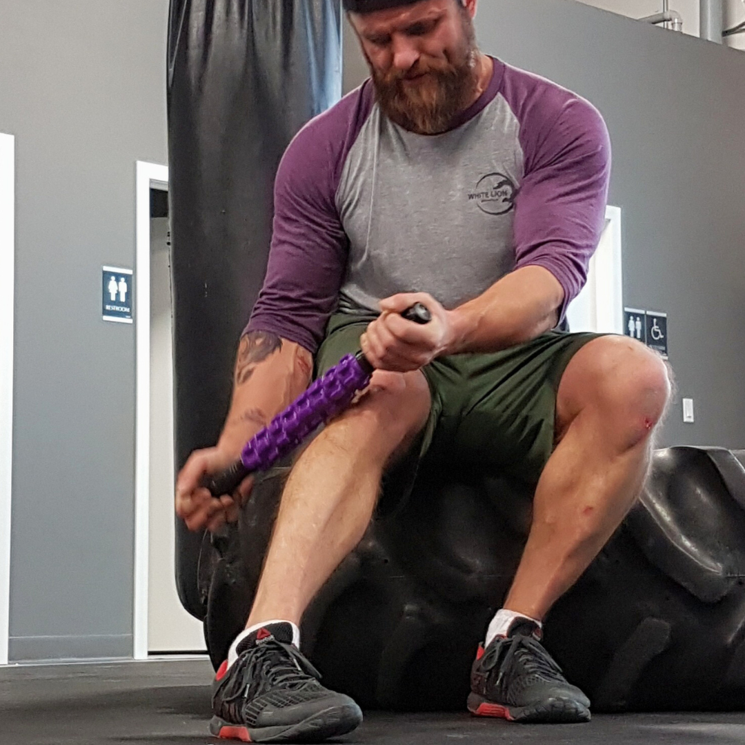 Man seated on large tractor tire inside of a gym use the White Lion Athletics Purple Rainmaker Massage Stick on his right, lower leg.