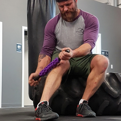 Man seated on large tractor tire inside of a gym use the White Lion Athletics Purple Rainmaker Massage Stick on his right, lower leg.
